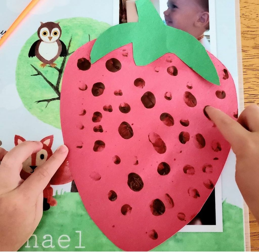A photo of a child finger painting red "seeds" onto a cut out picture of a red strawberry