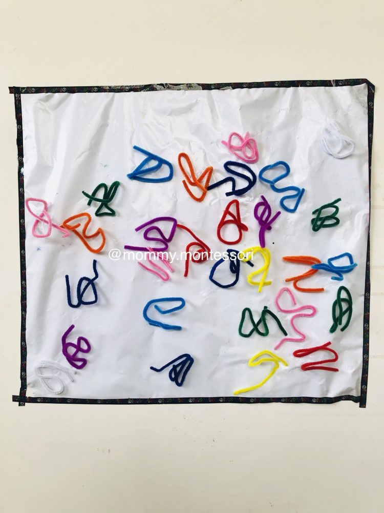 A photo of a white wall with a black framed white contact sheet. there are lots of brightly coloured pipe cleaners on top. they form lots of squiggly shapes.
