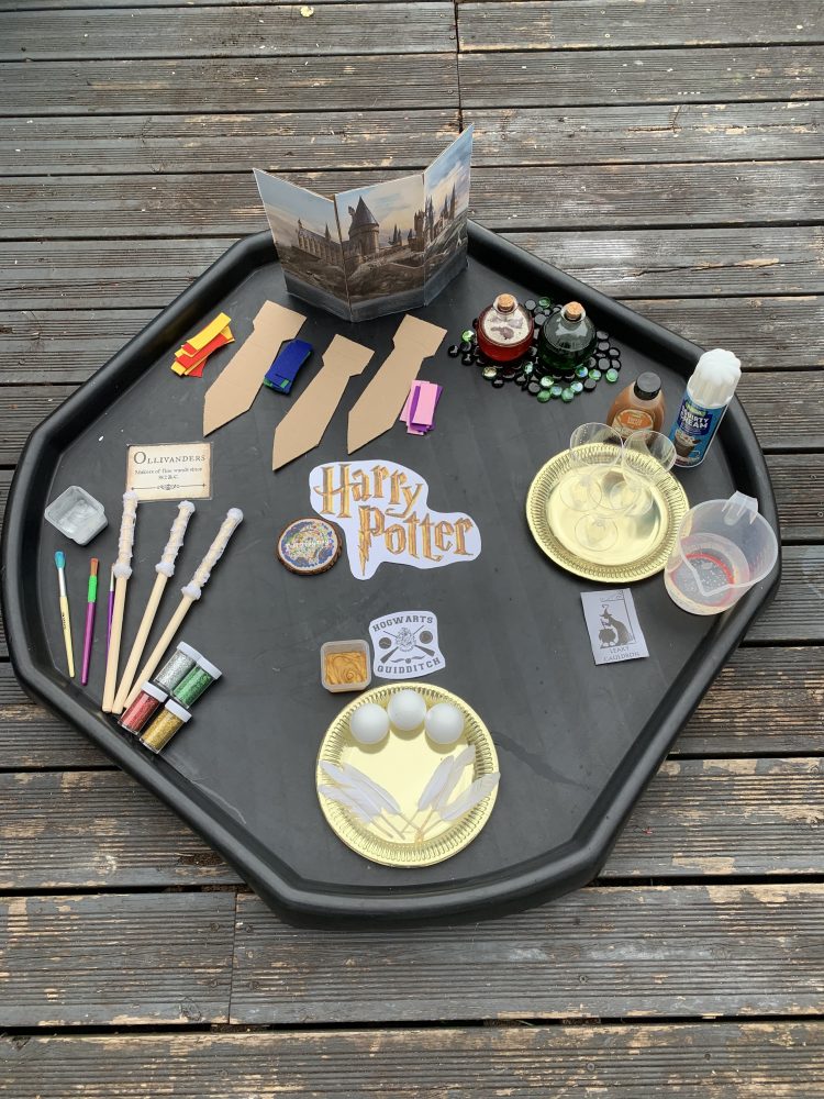 A photo of a tuff tray with Harry Potter written on,. there are craft items on it- including white balls and feathers on a gold plate.  there are brown cardboard ties.