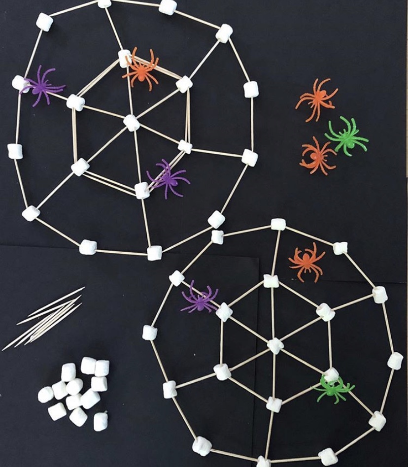 A photo of a back background with white marshmallows joined together with cocktail sticks to make a spideweb shape.  there are also colourful plastic spiders