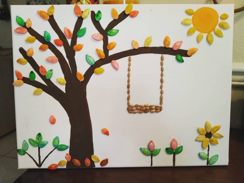 A photo of a white canvas with a brown tree, with colourful pumpkin seed "leaves", a swing, flowers and a sun