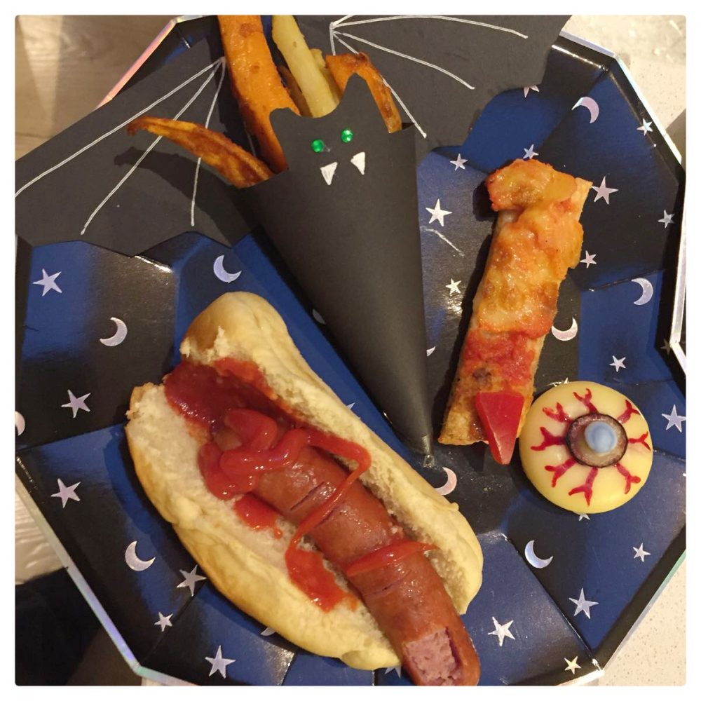 a hotdog, a black bat paper cone with chips in, a slice of pizza shaped like a witches finger and an eyeball on a paper plate with moons and stars on
