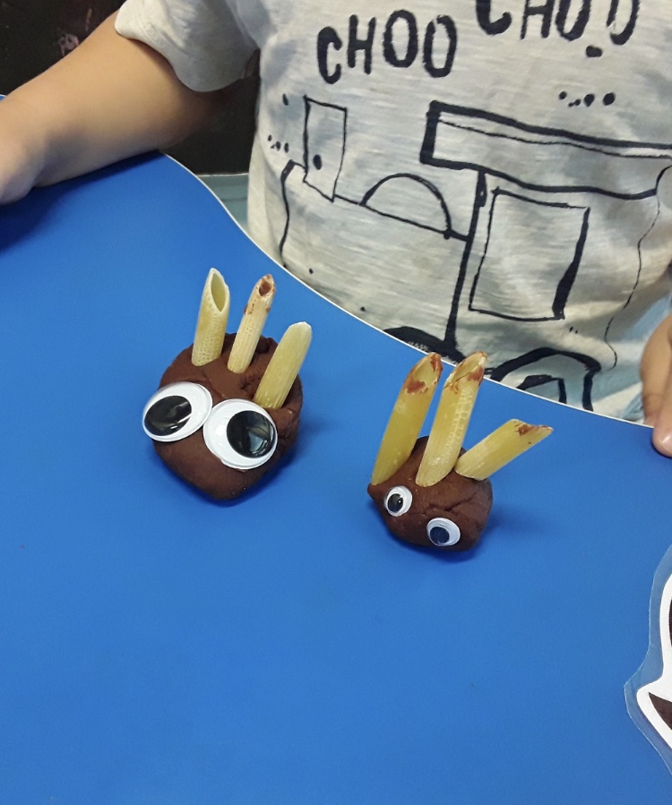 A photo of 2 brown playdoh hedghogs  both have googly eyes and 3 pasta shapes for spikes.  they are on a blue table.