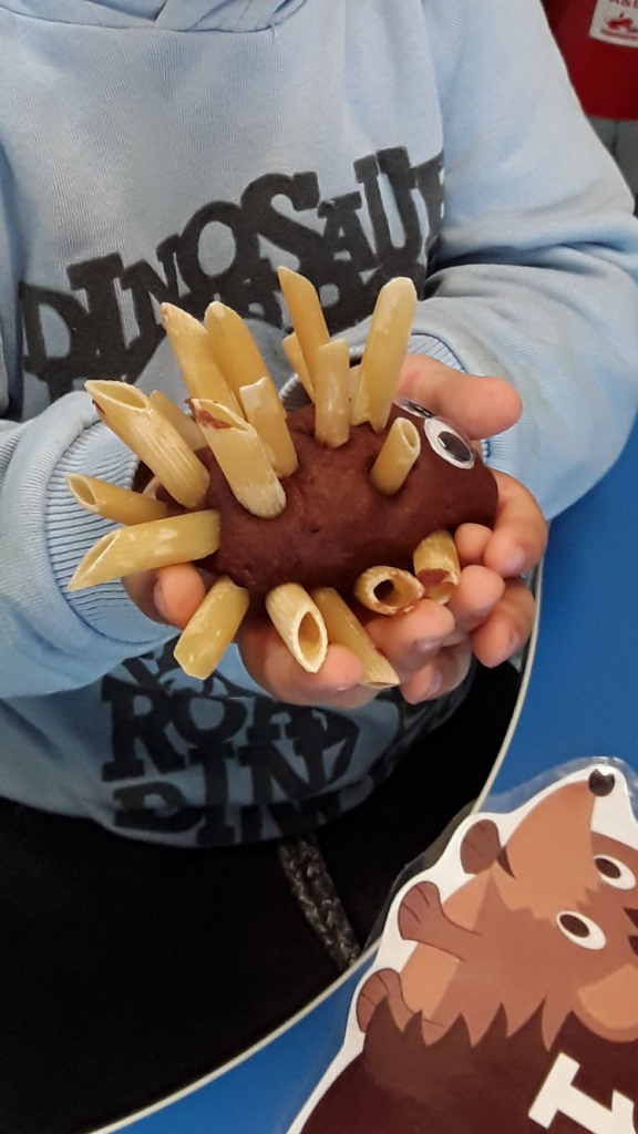 A photo of a child in a grey hoodie holding a brown playdoh hedgehog in his hands.  it had pasta shapes that act as its spikes. 