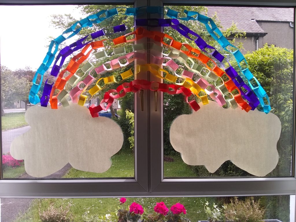 a photo of outside a window. on the window are coloured paper chains in the shape of a rainbow with a white cloud at either end