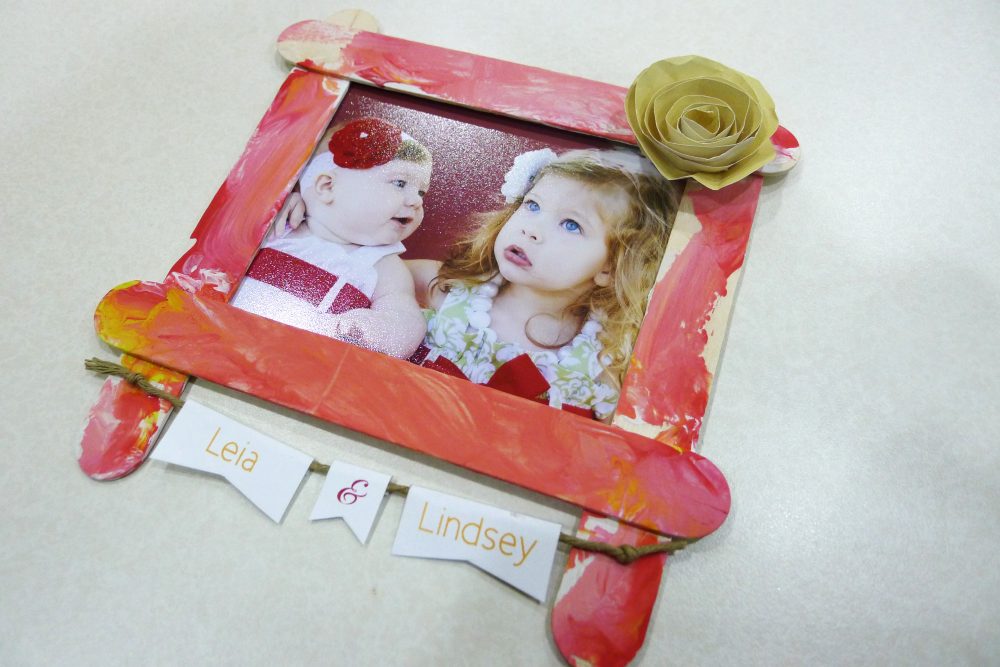 a photo of a red/pink frame with a yellow paper flower on  inside is a  photo of 2 girls