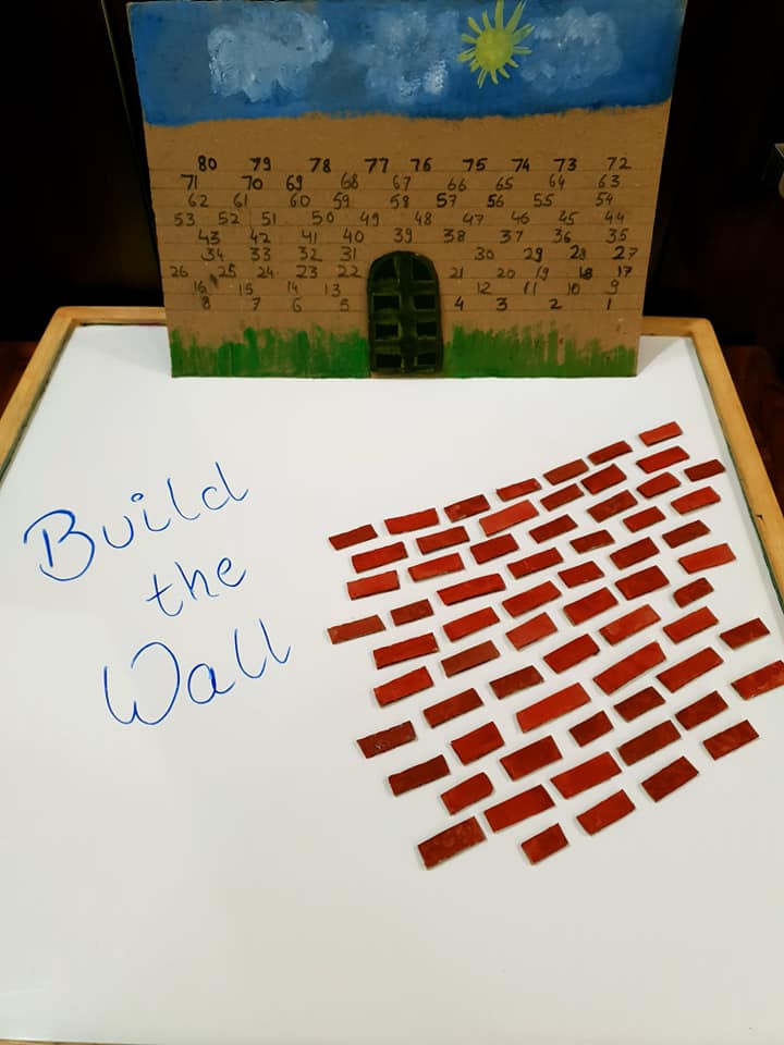 A photo of a whiteboard with the words build the wall written on.  Next to it are lots of mini red/brown bricks.  standing upright is a card board wall with numbers on