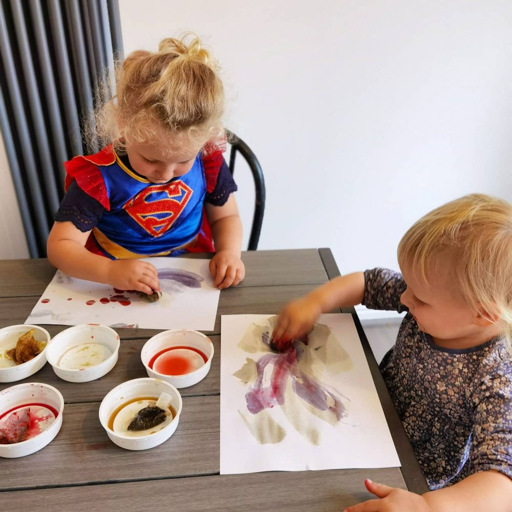 A photo of 2 children sitting at a table painting.  there are 5 bowls with coloured teabags inside