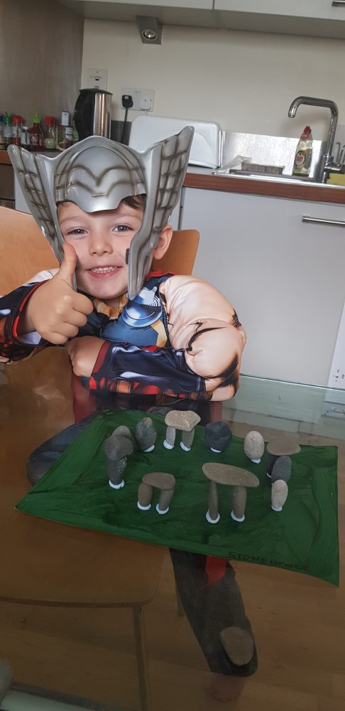 A photo of a boy in a costume smiling. thumb up.  there are pebbles on blu tac on green cardboard.