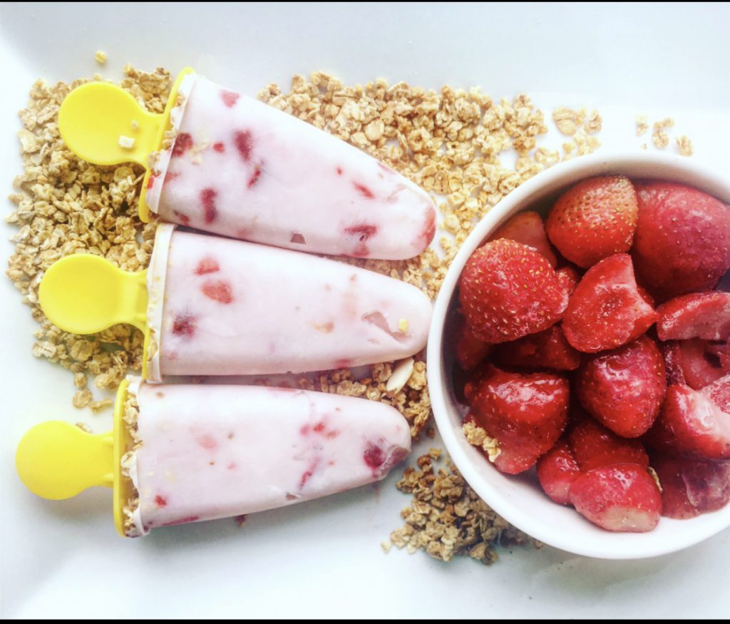 A photo of 2 pink yogurt parfait lollies- with yellow handles.  There is also a bowl of strawberries and some granola.  