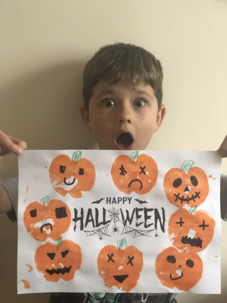 A photo of a boy holding a white piece of paper, with orange painted pumpkins.  it says Happy Halloween.