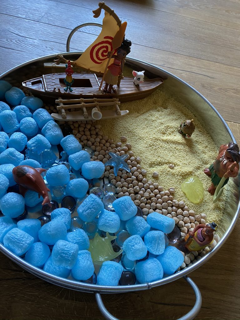 A photo of a silver tray with Moana plastic toy figure, a boat, blue plastic balls and sand and stones.