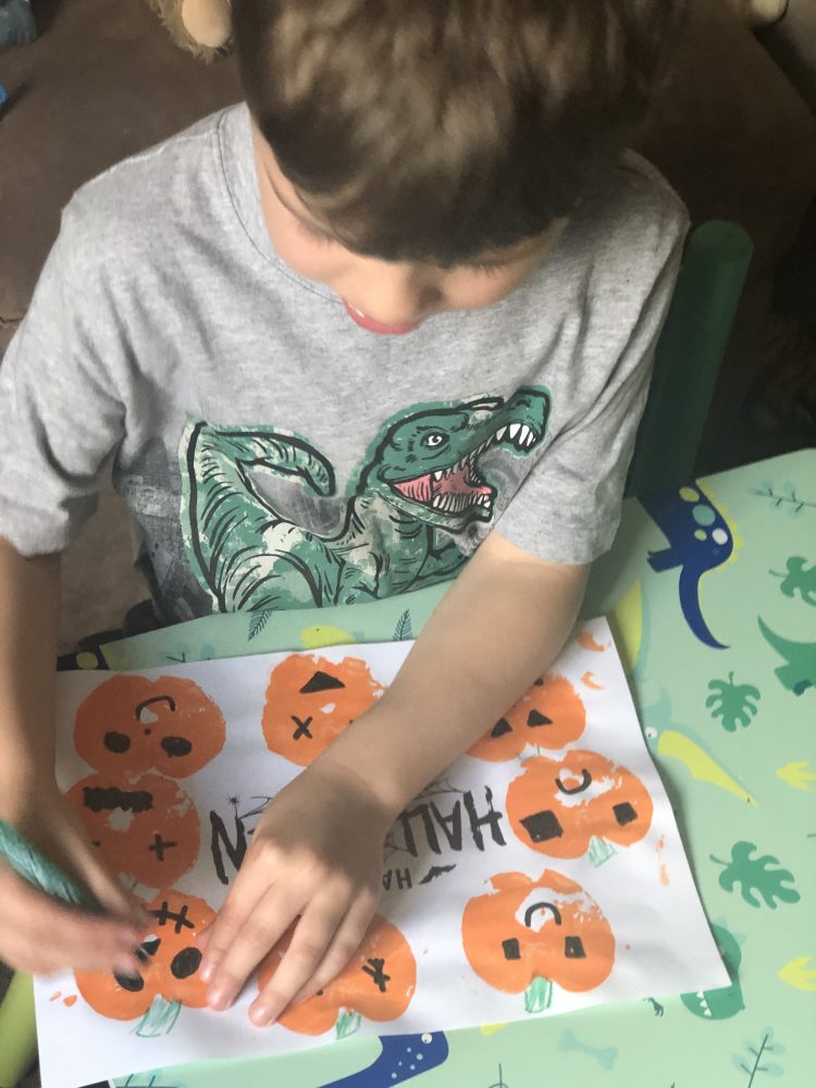 A photo of a boy drawing with green pen on a white piece of paper, with orange painted pumpkins