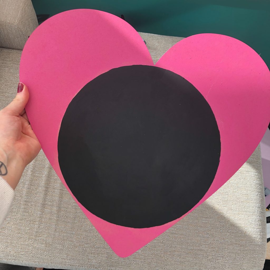 A photo of a hand holding a pink heart with a black circle painted on it.