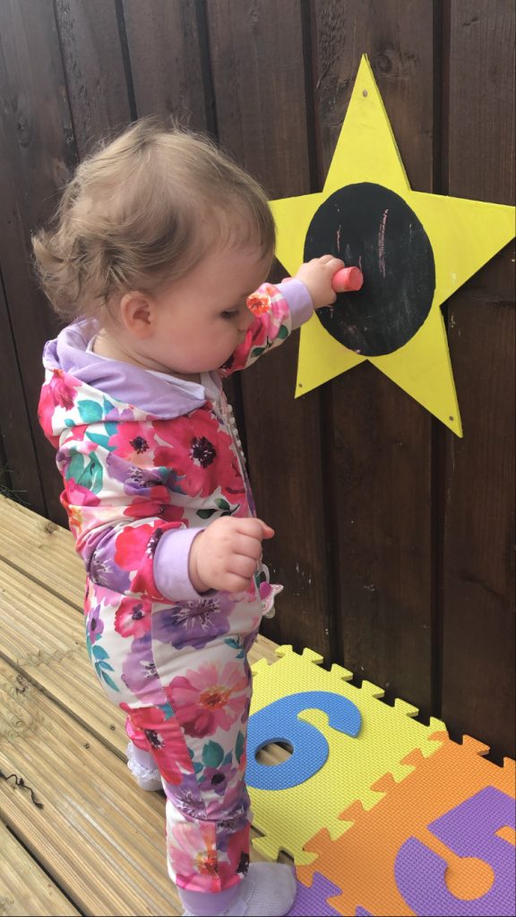 A photo of a little girl with a red chalk drawing on a yellow star , with a black circle... on a brown fence