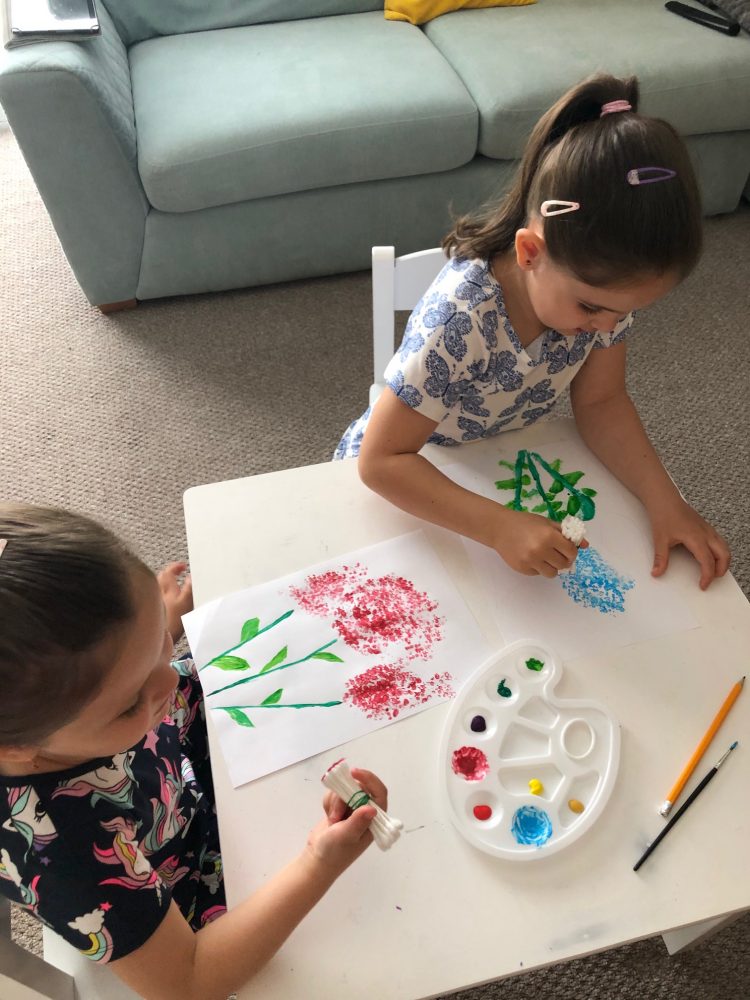 A photo of 2 girls at a white table, using cotton buds and paint to make flower pictures.
