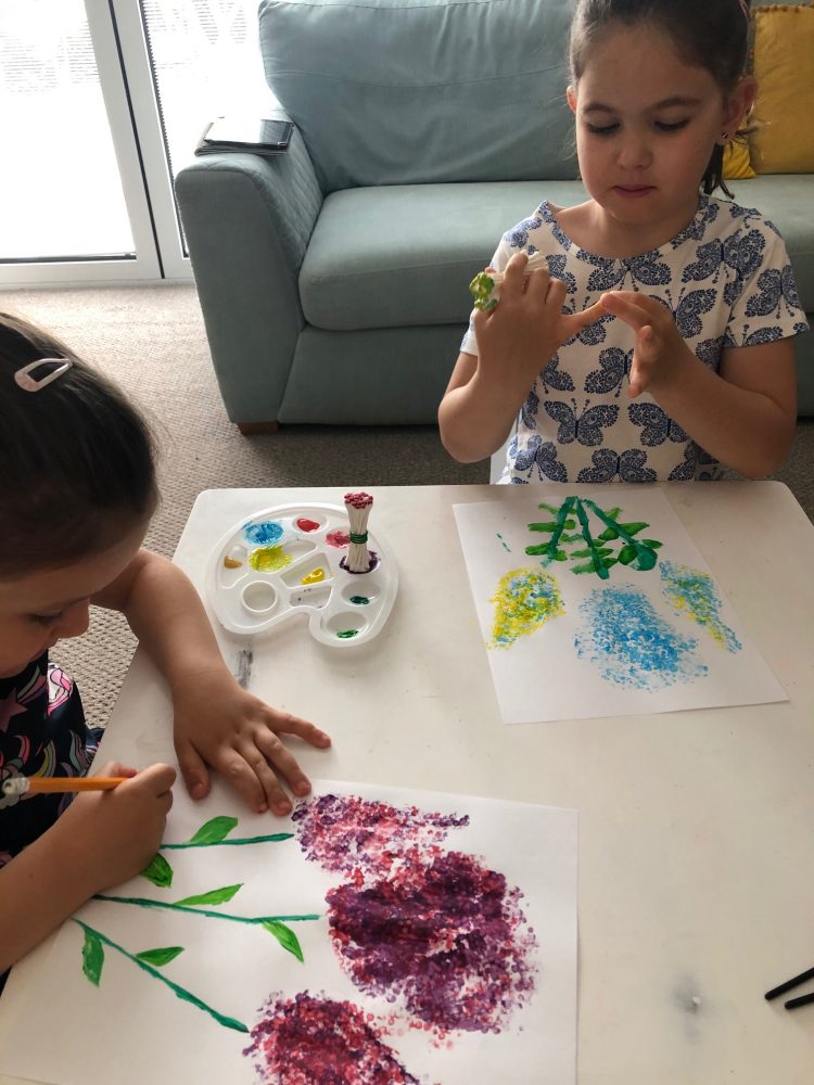 A photo of 2 girls at a white table, using cotton buds and paint to make flower pictures.