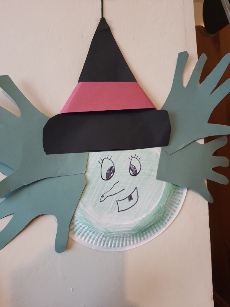 A photo of a paper plate witch craft.  with green paper hand cut outs, a black hat with a red stripe. 