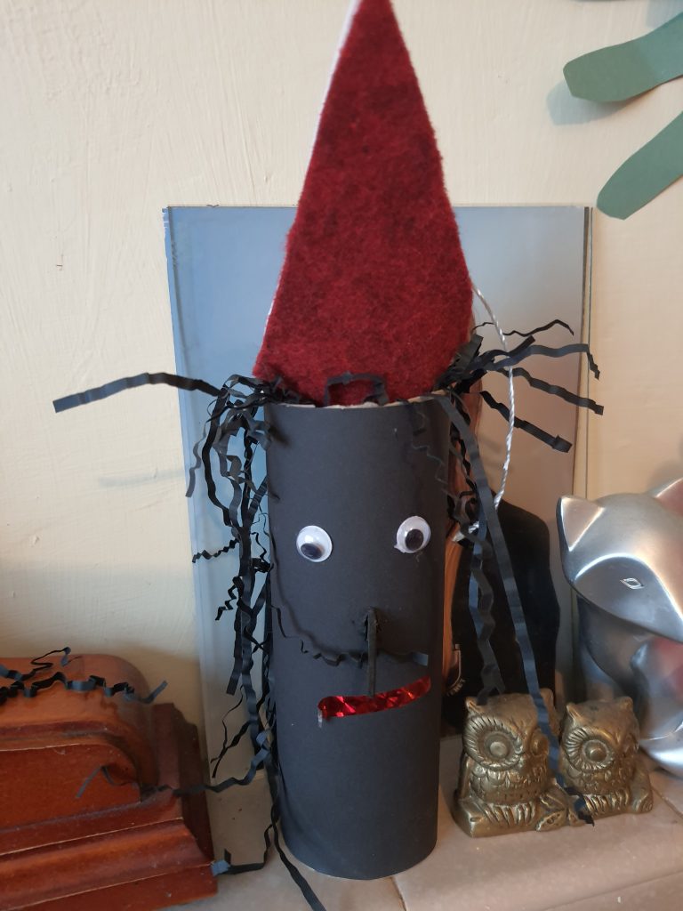 A photo of a black cardboard tube with black paper hair, a red felt hat and a face with googly eyes.  