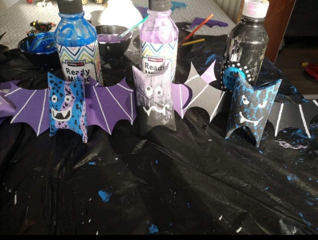 A photo of 3 home made cardboard bats, coloured blue, burple, black and white.  there are three bottles of paint on the table too. 