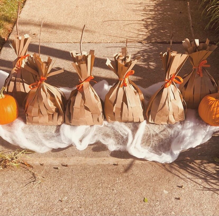 A photo of 6 brown paper bags tied with orange ribbon.  there are also 2 pumpkins on top of white fluffy material on a path