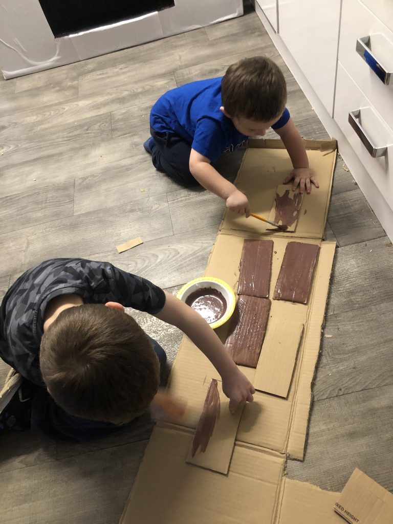 A photo of 2 boys using brown paint to paint cardboard bricks 