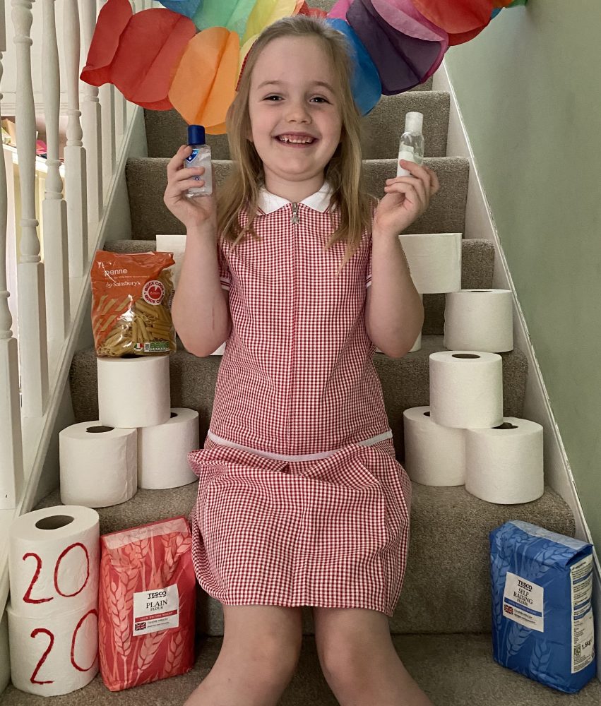 A photo of a girl in a red and white school summer dress.  she is holding hand sanitizer and has lots of toilet rolls and flour packets around her.