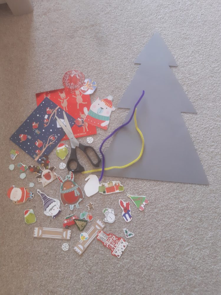 A photo of a cardboard tree on the floor with Christmas themed cut out paper and scissors next to it.