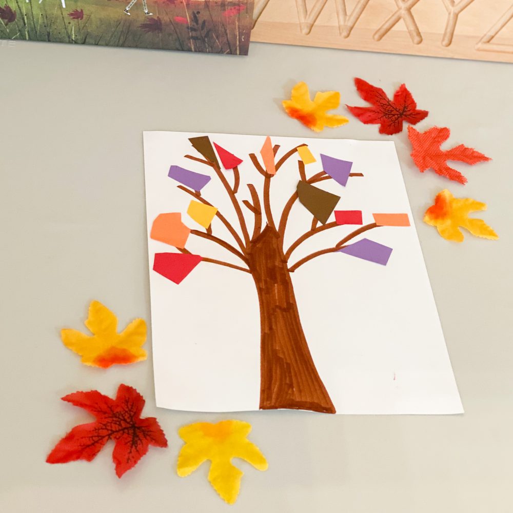 A photo of a book and a piece of paper with a tree drawn on, there is also coloured paper and autumn colour leaves